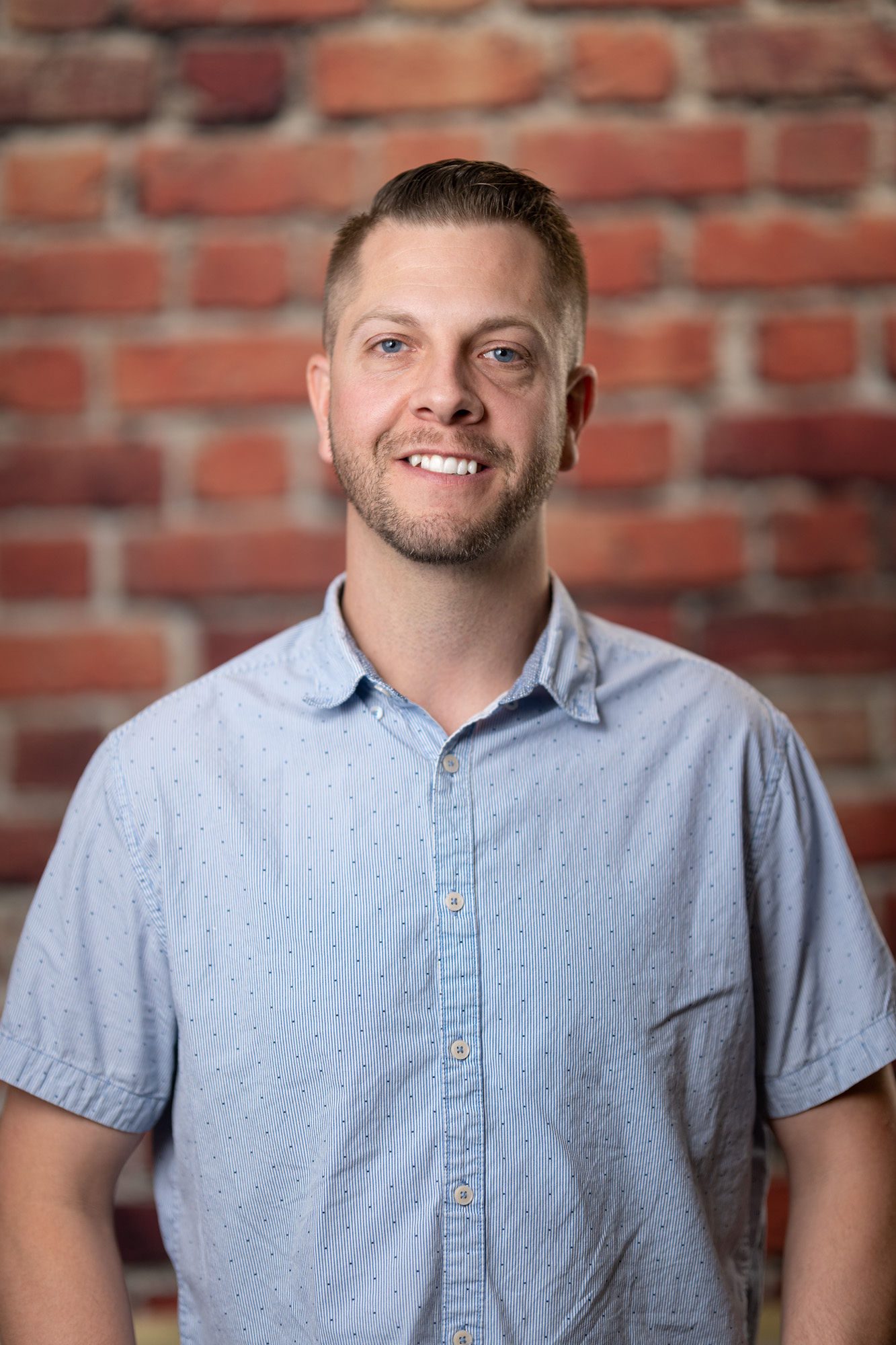 Josh Cornwell A man with short hair and a trimmed beard wearing a light blue, short-sleeved button-up shirt stands against a background of red brick.