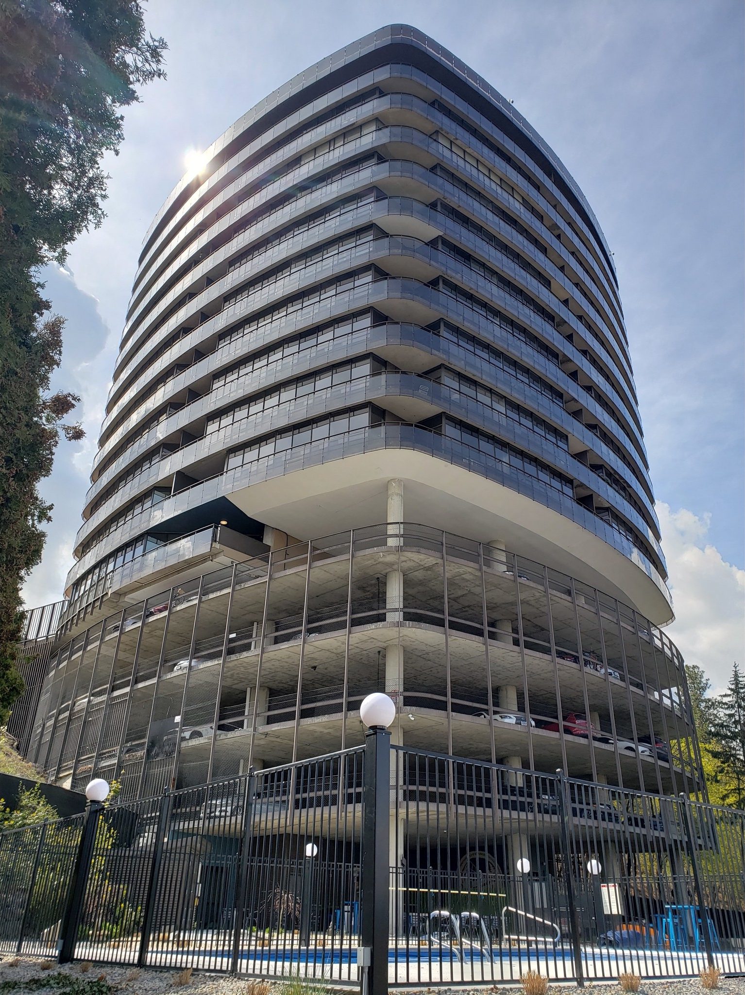 Riverfalls Tower A tall, multi-story modern building with glass exterior and an open ground level, surrounded by a black metal fence with white spherical lights. Trees and blue sky are in the background.